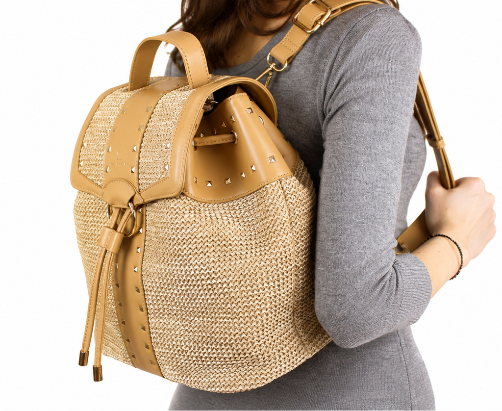 A woven straw backpack with tan leather trim, studded details, and a top handle, shown against a plain white background.